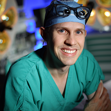 Gedge Rosson poses for a photo in an operating room wearing green scrubs and a light blue surgical cap